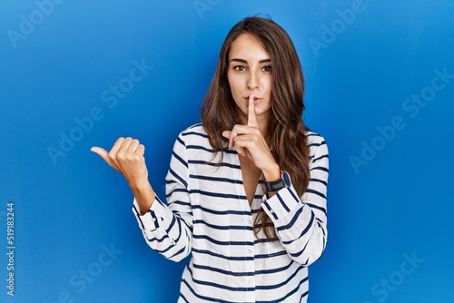 Young hispanic woman standing over blue isolated background asking to be quiet with finger on lips pointing with hand to the side. silence and secret concept. © Krakenimages.com