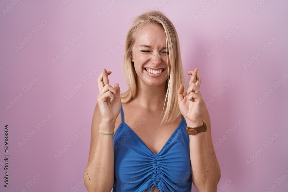 Young caucasian woman standing over pink background gesturing finger crossed smiling with hope and eyes closed. luck and superstitious concept. Young caucasian woman standing over pink background gesturing finger crossed smiling with hope and eyes closed. luck and superstitious concept.