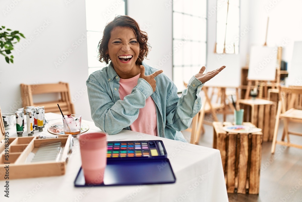 Middle age hispanic woman at art studio amazed and smiling to the camera while presenting with hand and pointing with finger. Middle age hispanic woman at art studio amazed and smiling to the camera while presenting with hand and pointing with finger.