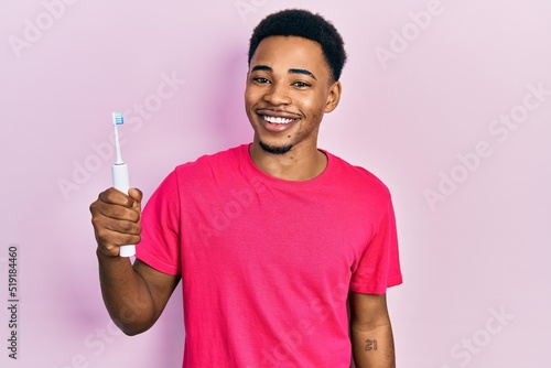 Young african american man holding electric toothbrush looking positive and happy standing and smiling with a confident smile showing teeth