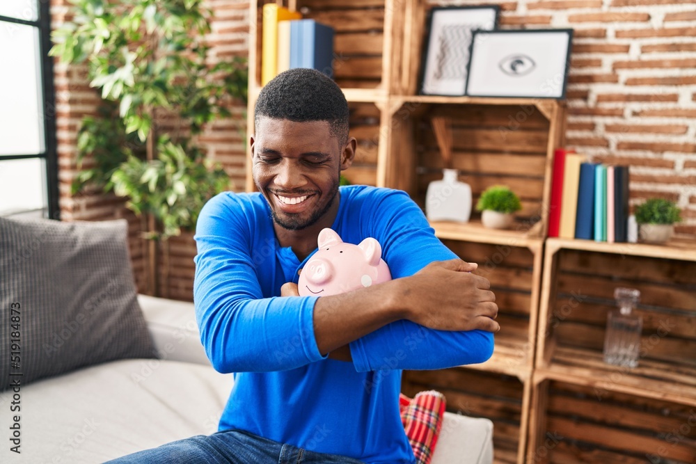 Obraz premium Young african american man hugging piggy bank sitting on sofa at home