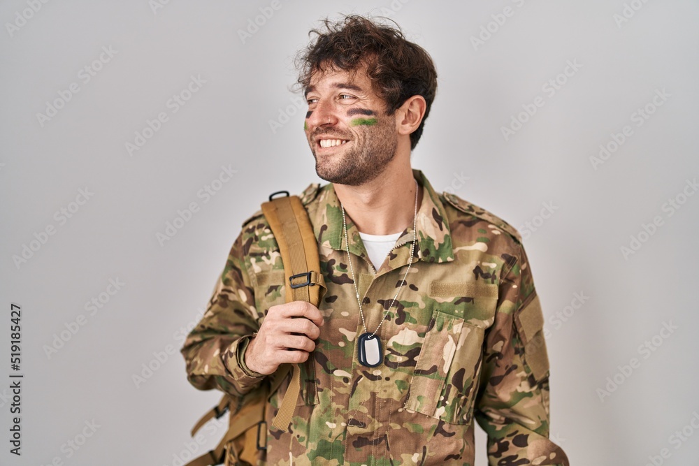 Hispanic young man wearing camouflage army uniform looking away to side with smile on face, natural expression. laughing confident.