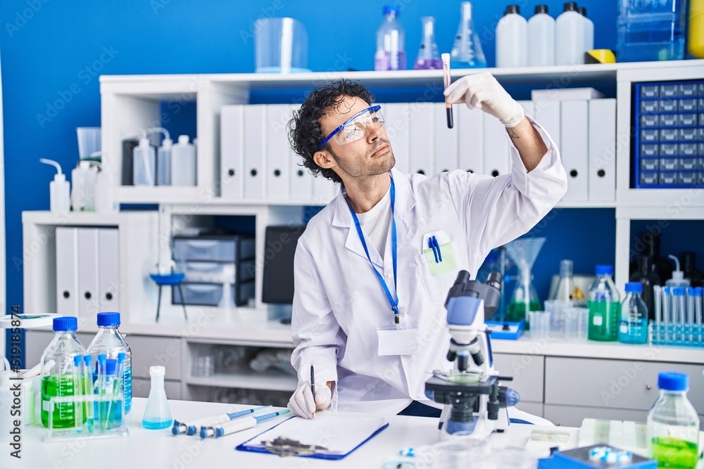 Fototapeta premium Young hispanic man scientist writing on document holding blood test tube at laboratory