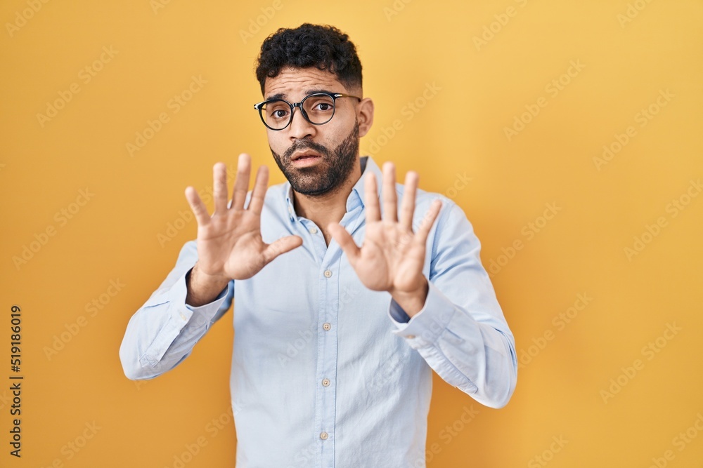 Hispanic man with beard standing over yellow background afraid and terrified with fear expression stop gesture with hands, shouting in shock. panic concept. Hispanic man with beard standing over yellow background afraid and terrified with fear expression stop gesture with hands, shouting in shock. panic concept.