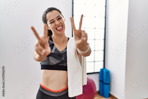 Young brunette woman wearing sportswear and towel at the gym smiling with tongue out showing fingers of both hands doing victory sign. number two.