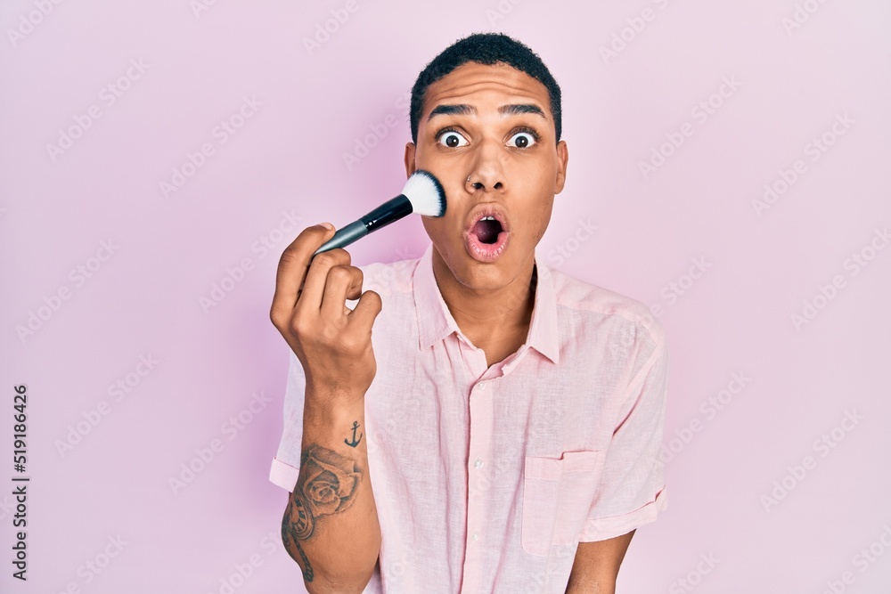 Young african american guy holding makeup brush scared and amazed with open mouth for surprise, disbelief face Young african american guy holding makeup brush scared and amazed with open mouth for surprise, disbelief face