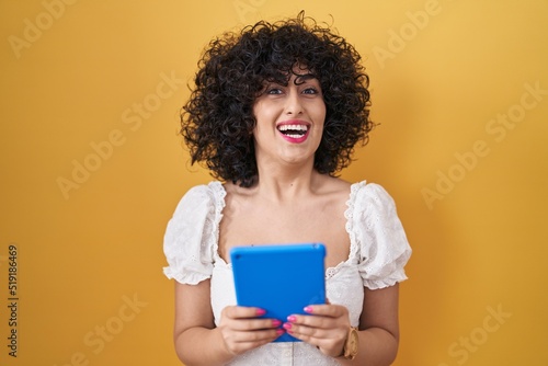 Young brunette woman with curly hair using touchpad over yellow background celebrating crazy and amazed for success with open eyes screaming excited.