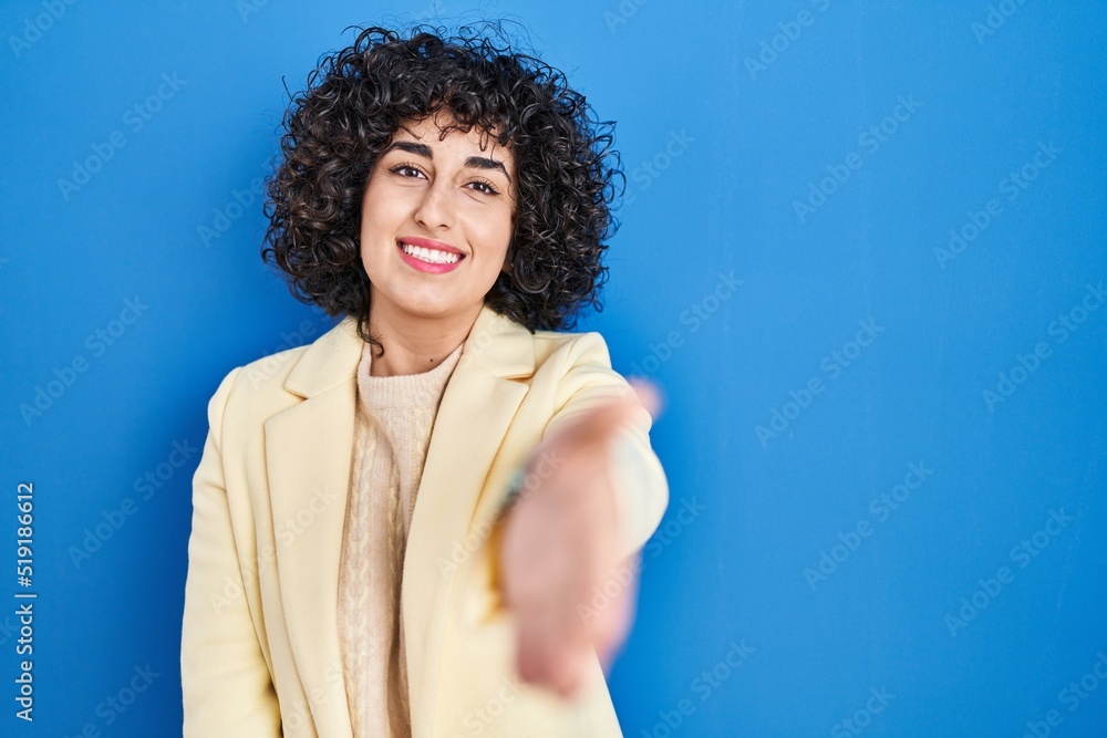 Young brunette woman with curly hair standing over blue background smiling friendly offering handshake as greeting and welcoming. successful business. Young brunette woman with curly hair standing over blue background smiling friendly offering handshake as greeting and welcoming. successful business.