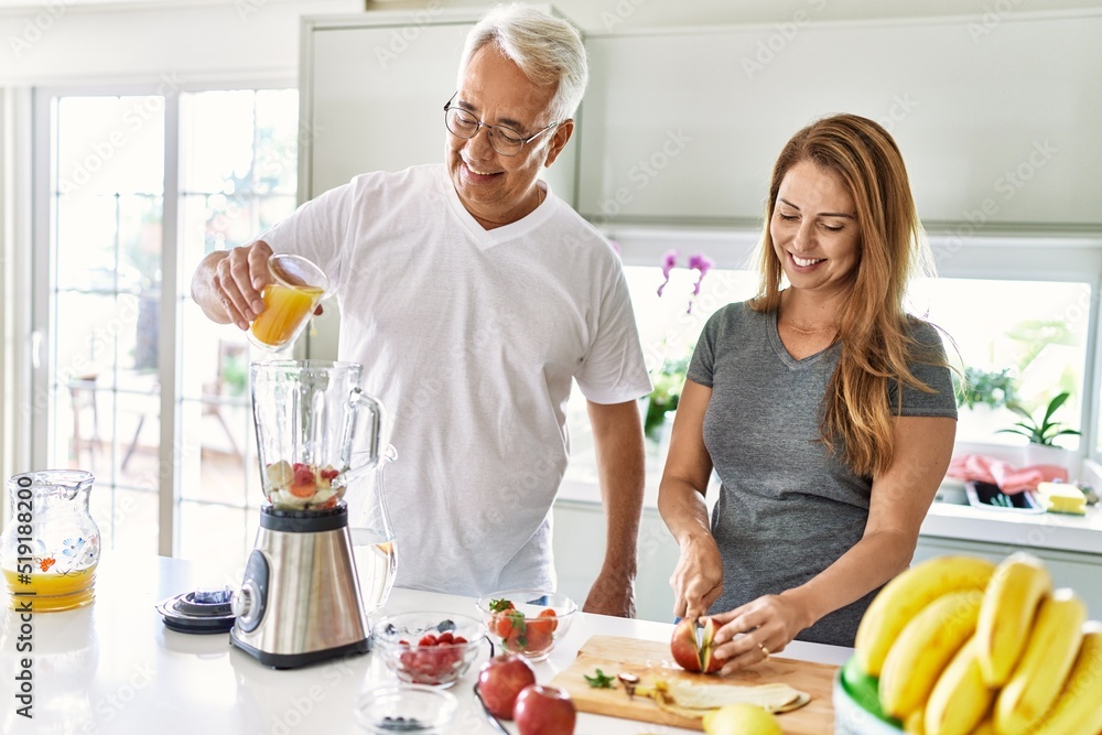 Middle age hispanic couple pouring juice on mixer machine cooking smoothie at the kitchen. Middle age hispanic couple pouring juice on mixer machine cooking smoothie at the kitchen.