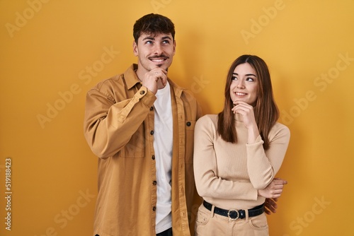 Young hispanic couple standing over yellow background with hand on chin thinking about question, pensive expression. smiling and thoughtful face. doubt concept.