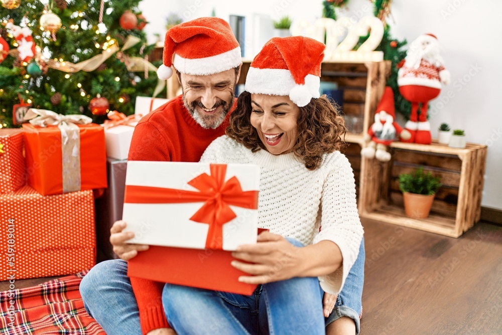 Middle age hispanic couple wearing christmas hat. Sitting on the floor opening gift at home. Middle age hispanic couple wearing christmas hat. Sitting on the floor opening gift at home.