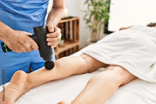 Middle age man and woman wearing therapist uniform having leg massage session using percussion pistol at beauty center