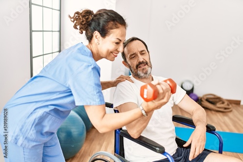 Middle age man and woman having rehab session using dumbbell sitting on wheelchair at physiotherapy clinic