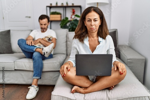 Hispanic middle age couple at home, woman using laptop skeptic and nervous, frowning upset because of problem. negative person. © Krakenimages.com