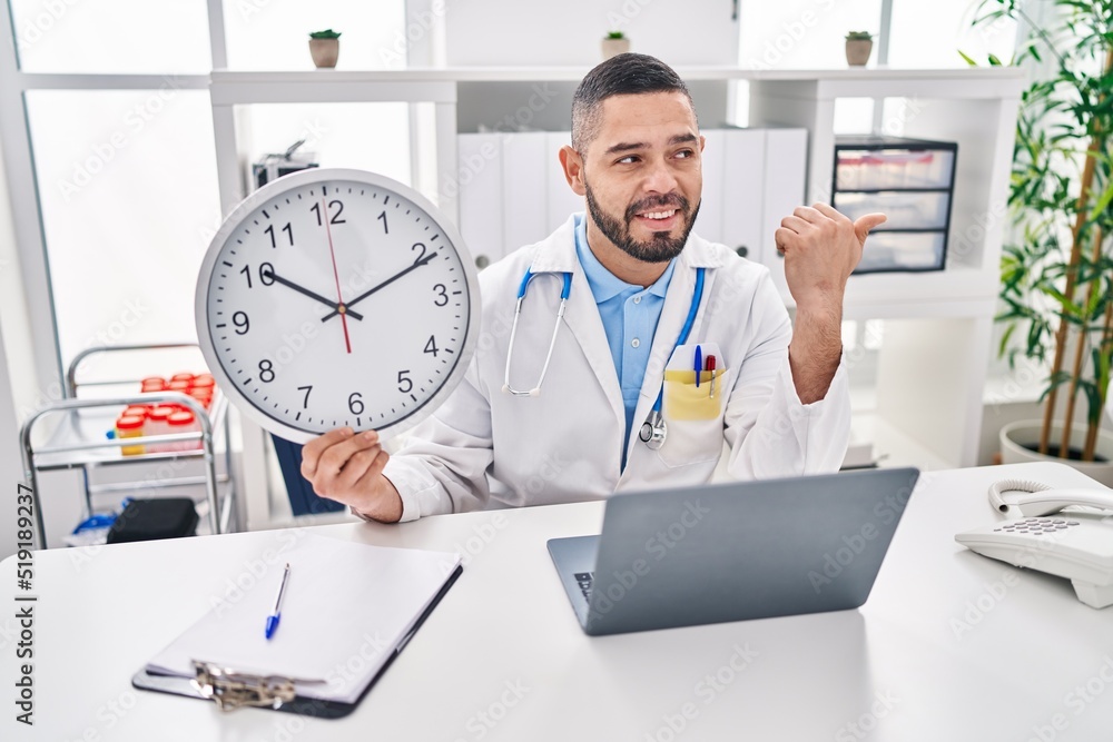 Hispanic doctor man holding clock at the clinic pointing thumb up to the side smiling happy with open mouth Hispanic doctor man holding clock at the clinic pointing thumb up to the side smiling happy with open mouth