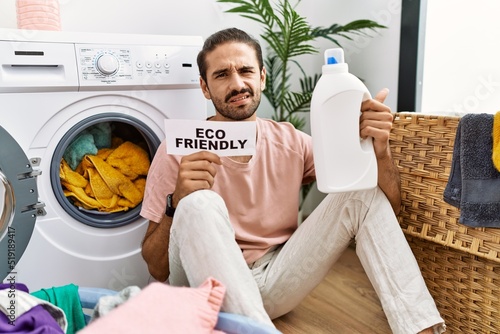 Fotografia Young hispanic man doing laundry holding eco friendly paper clueless and confused expression