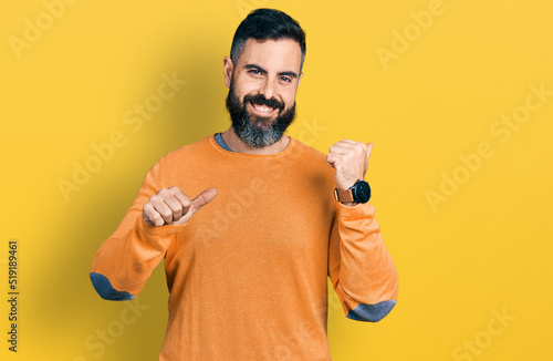 Hispanic man with beard wearing casual winter sweater pointing to the back behind with hand and thumbs up, smiling confident
