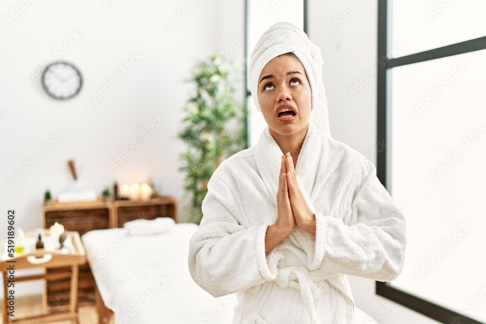 Young brunette woman wearing towel and bathrobe standing at beauty center begging and praying with hands together with hope expression on face very emotional and worried. begging. Young brunette woman wearing towel and bathrobe standing at beauty center begging and praying with hands together with hope expression on face very emotional and worried. begging.