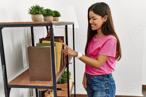 Young latin woman smiling confident choosing book of shelving at home
