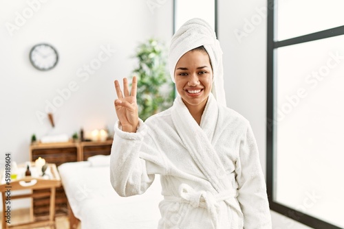 Young brunette woman wearing towel and bathrobe standing at beauty center showing and pointing up with fingers number three while smiling confident and happy.