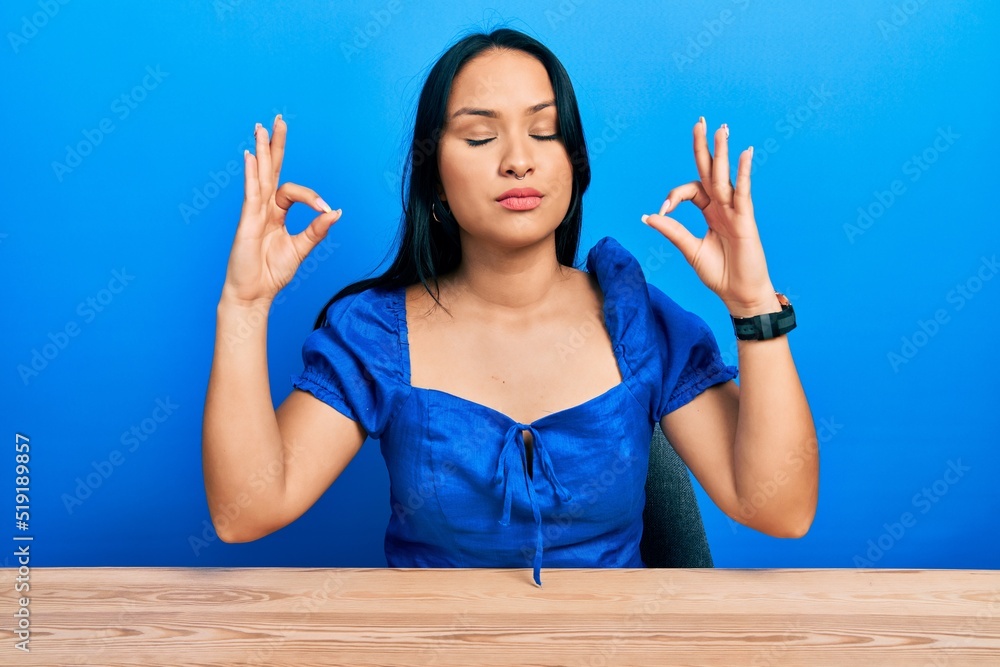 Beautiful hispanic woman with nose piercing sitting on the table relax and smiling with eyes closed doing meditation gesture with fingers. yoga concept. Beautiful hispanic woman with nose piercing sitting on the table relax and smiling with eyes closed doing meditation gesture with fingers. yoga concept.