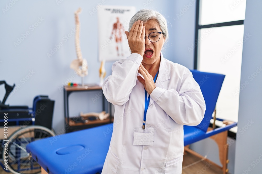 Middle age woman with grey hair working at pain recovery clinic yawning tired covering half face, eye and mouth with hand. face hurts in pain. Middle age woman with grey hair working at pain recovery clinic yawning tired covering half face, eye and mouth with hand. face hurts in pain.
