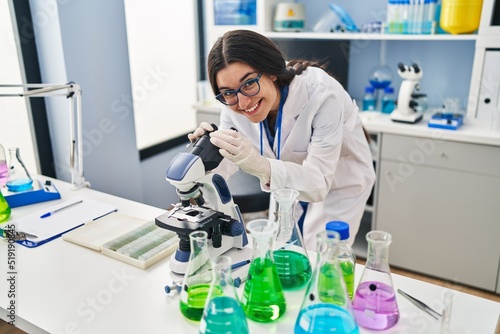 Young hispanic woman wearing scientist uniform using microscope at laboratory