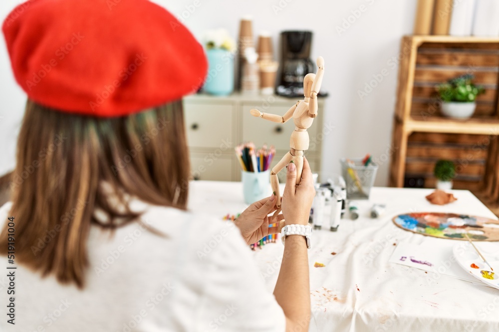 Young hispanic artist woman on back view wearing french beret holding manikin at art studio.