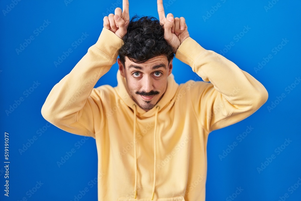 Hispanic man standing over blue background doing funny gesture with finger over head as bull horns Hispanic man standing over blue background doing funny gesture with finger over head as bull horns