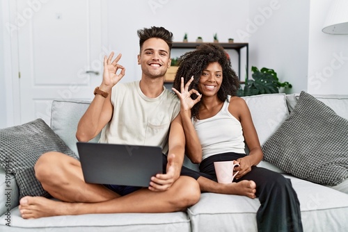 Young interracial couple using laptop at home sitting on the sofa smiling positive doing ok sign with hand and fingers. successful expression.