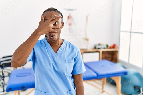 Black woman with braids working at pain recovery clinic peeking in shock covering face and eyes with hand, looking through fingers with embarrassed expression.