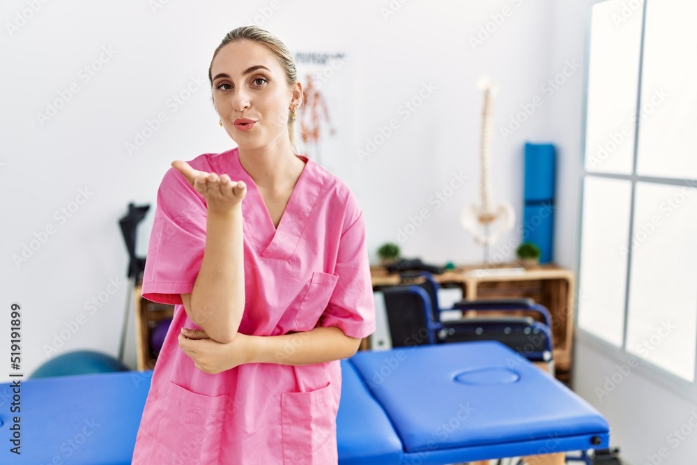 Young blonde woman working at pain recovery clinic looking at the camera blowing a kiss with hand on air being lovely and sexy. love expression. Young blonde woman working at pain recovery clinic looking at the camera blowing a kiss with hand on air being lovely and sexy. love expression.