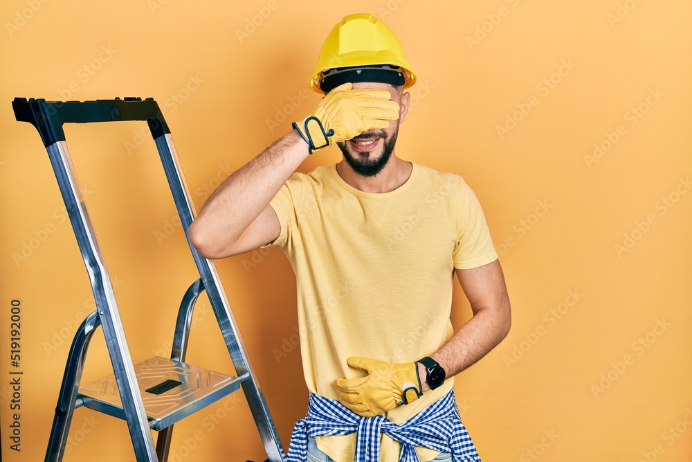 Handsome man with beard by construction stairs wearing hardhat smiling and laughing with hand on face covering eyes for surprise. blind concept. Handsome man with beard by construction stairs wearing hardhat smiling and laughing with hand on face covering eyes for surprise. blind concept.