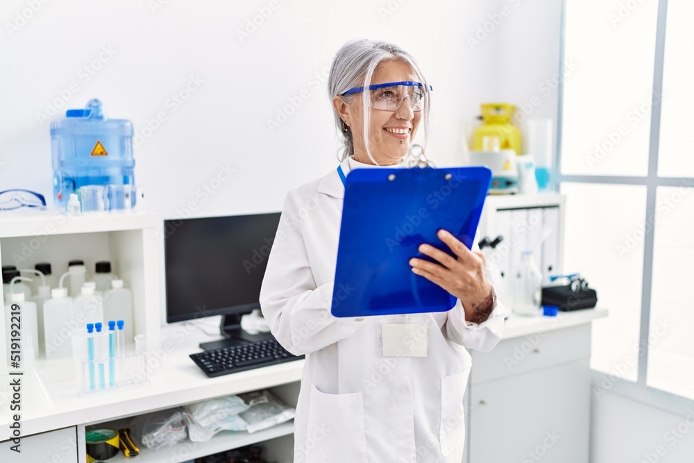 Middle age grey-haired woman wearing scientist uniform writing on clipboard working at laboratory Middle age grey-haired woman wearing scientist uniform writing on clipboard working at laboratory