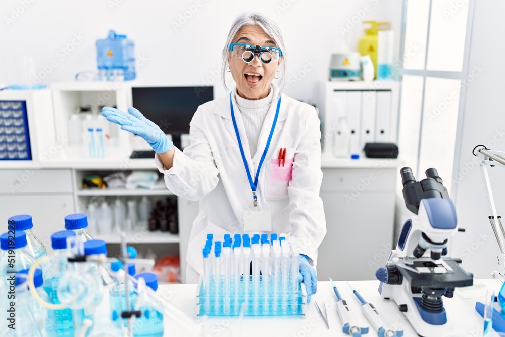 Middle age grey-haired woman wearing scientist uniform celebrating achievement with happy smile and winner expression with raised hand Middle age grey-haired woman wearing scientist uniform celebrating achievement with happy smile and winner expression with raised hand