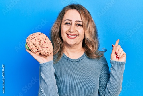 Middle age caucasian woman holding brain smiling happy pointing with hand and finger to the side