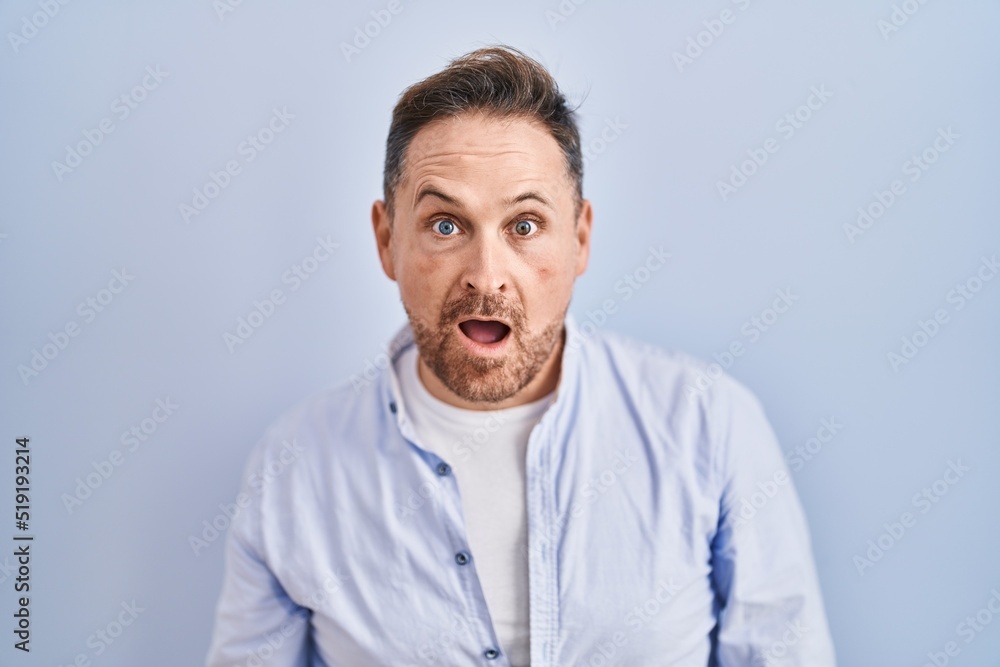 Middle age caucasian man standing over blue background afraid and shocked with surprise and amazed expression, fear and excited face.