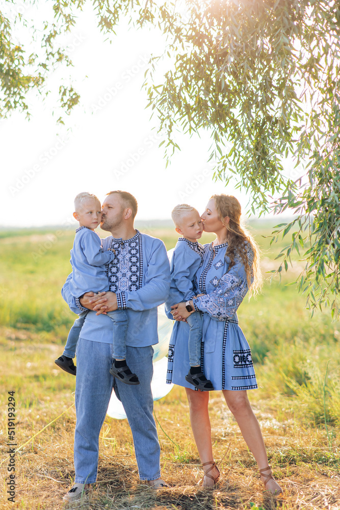 Fototapeta premium Happy parents hold their two twins sons on the hands and kiss them during walk on meadow.