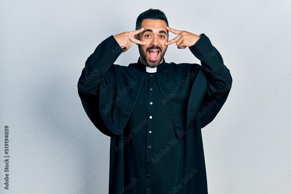 Handsome hispanic man with beard wearing catholic priest robe doing peace symbol with fingers over face, smiling cheerful showing victory Handsome hispanic man with beard wearing catholic priest robe doing peace symbol with fingers over face, smiling cheerful showing victory