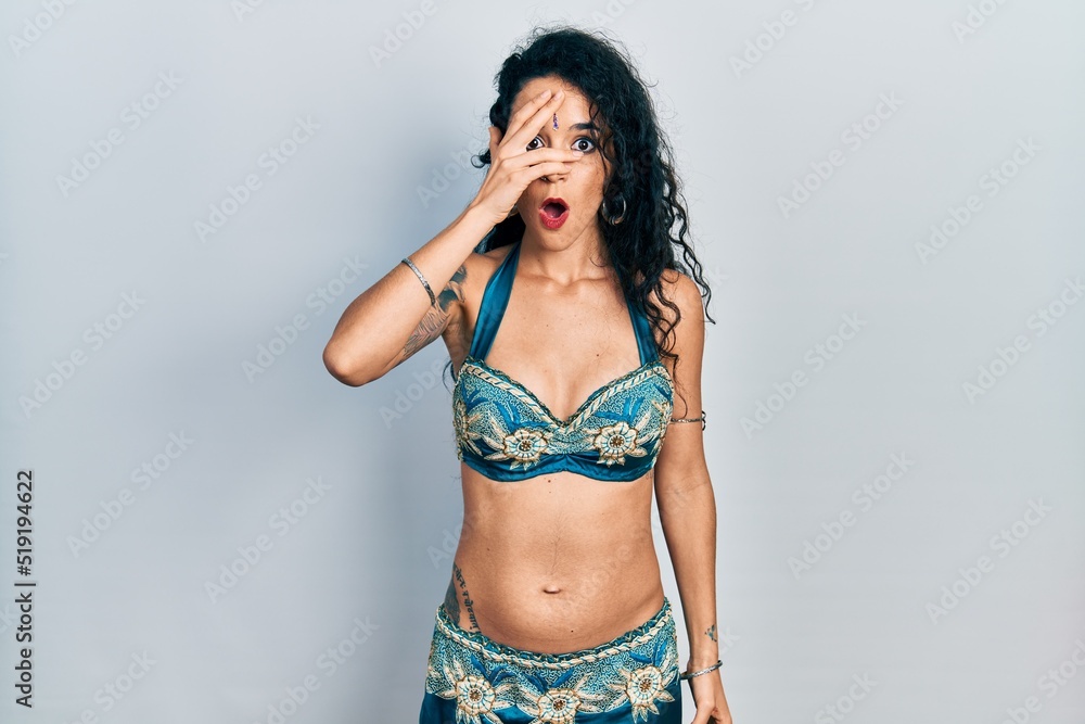 Young woman wearing bindi and traditional belly dance clothes peeking in shock covering face and eyes with hand, looking through fingers with embarrassed expression. Young woman wearing bindi and traditional belly dance clothes peeking in shock covering face and eyes with hand, looking through fingers with embarrassed expression.