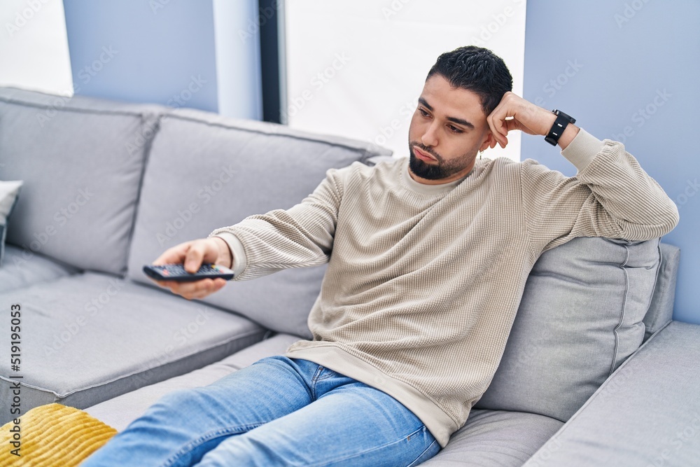 Young arab man watching tv with boring expression at home