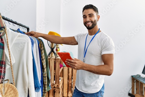 Young arab man shopkeeper using touchpad standing by clothes rack at clothing store