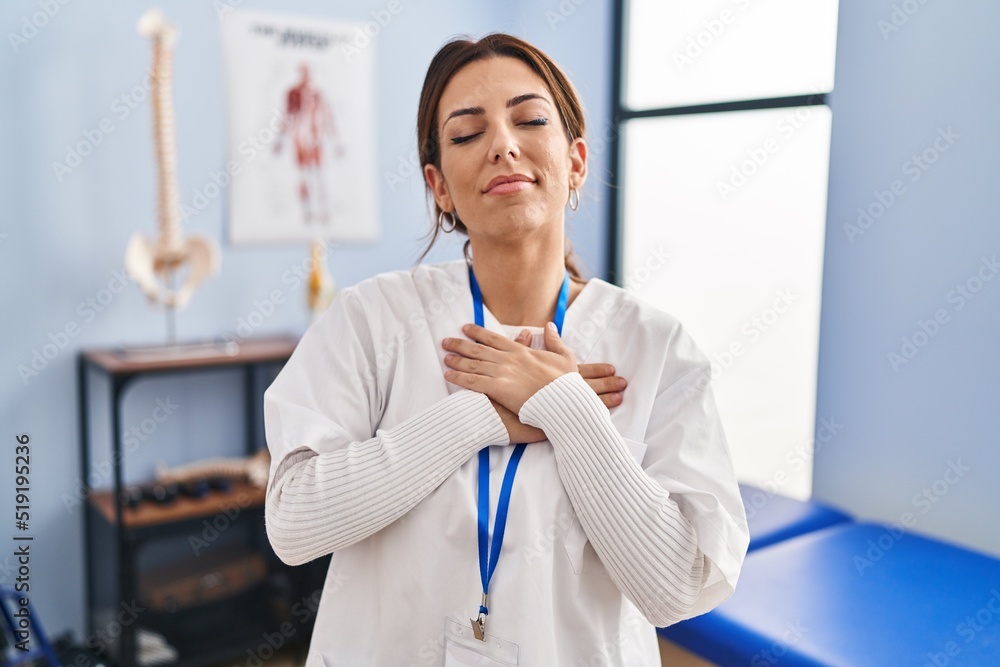Young brunette woman working at pain recovery clinic smiling with hands on chest with closed eyes and grateful gesture on face. health concept. Young brunette woman working at pain recovery clinic smiling with hands on chest with closed eyes and grateful gesture on face. health concept.