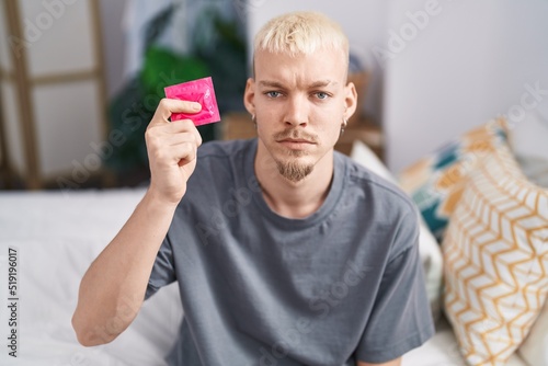 Young caucasian man holding condom sitting on bed thinking attitude and sober expression looking self confident