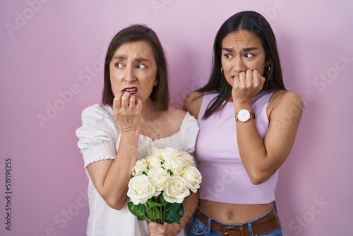 Hispanic mother and daughter holding bouquet of white flowers looking stressed and nervous with hands on mouth biting nails. anxiety problem.