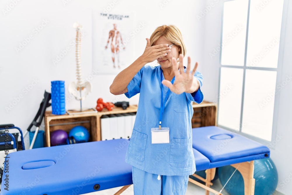 Middle age blonde woman working at pain recovery clinic covering eyes with hands and doing stop gesture with sad and fear expression. embarrassed and negative concept. Middle age blonde woman working at pain recovery clinic covering eyes with hands and doing stop gesture with sad and fear expression. embarrassed and negative concept.
