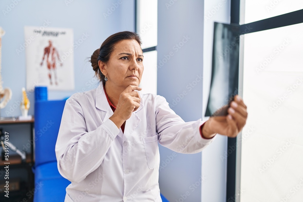 Fototapeta premium Middle age hispanic woman physiotherapist holding radiography at rehab clinic