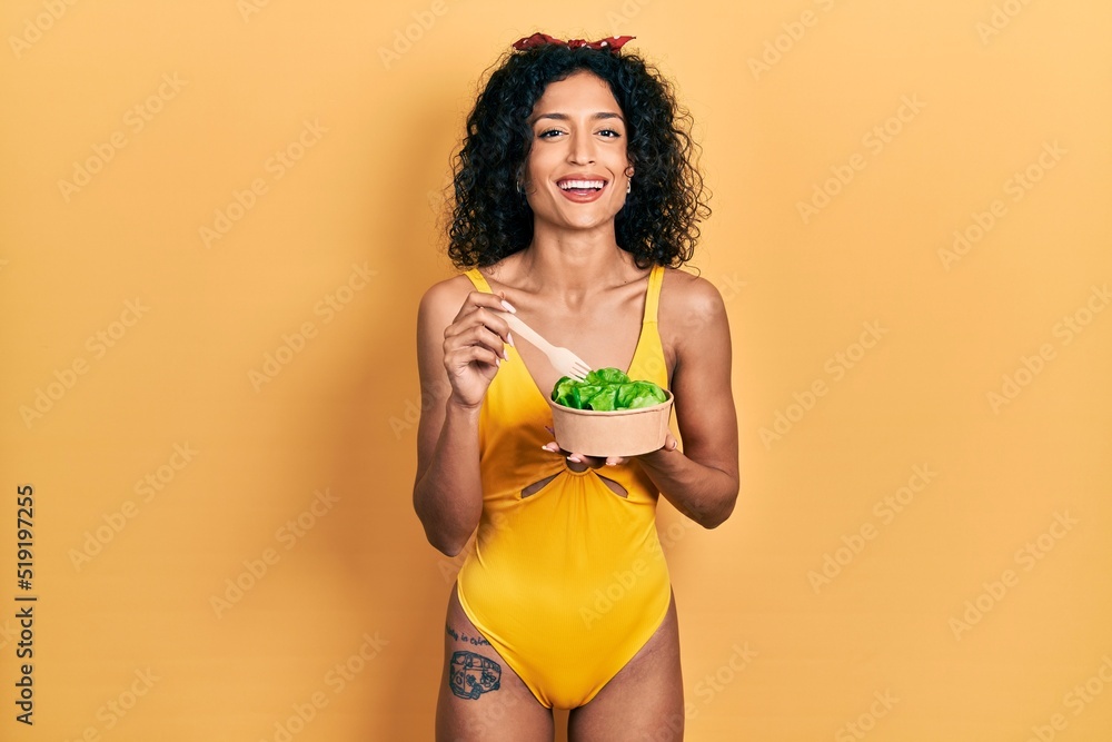 Naklejka premium Young latin girl wearing swimsuit eating salad smiling and laughing hard out loud because funny crazy joke.