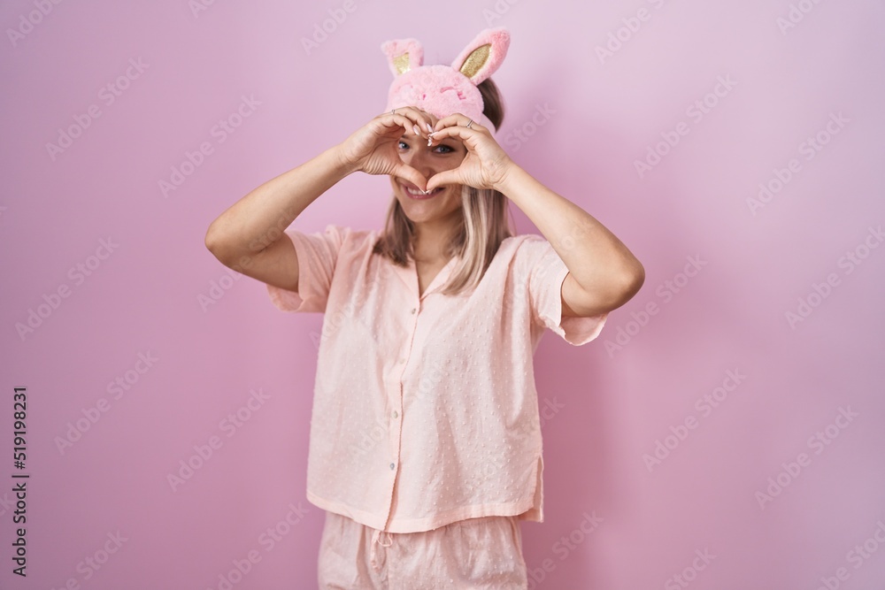 Blonde caucasian woman wearing sleep mask and pajama doing heart shape with hand and fingers smiling looking through sign