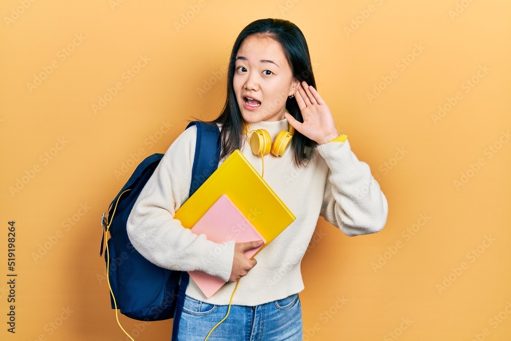 Young chinese girl holding student backpack and books smiling with hand over ear listening an hearing to rumor or gossip. deafness concept. Young chinese girl holding student backpack and books smiling with hand over ear listening an hearing to rumor or gossip. deafness concept.